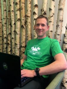 Ingo Steinke mit Laptop und grünem "Woodhack"-T-Shirt vor einer Wand aus Birkenstämmen