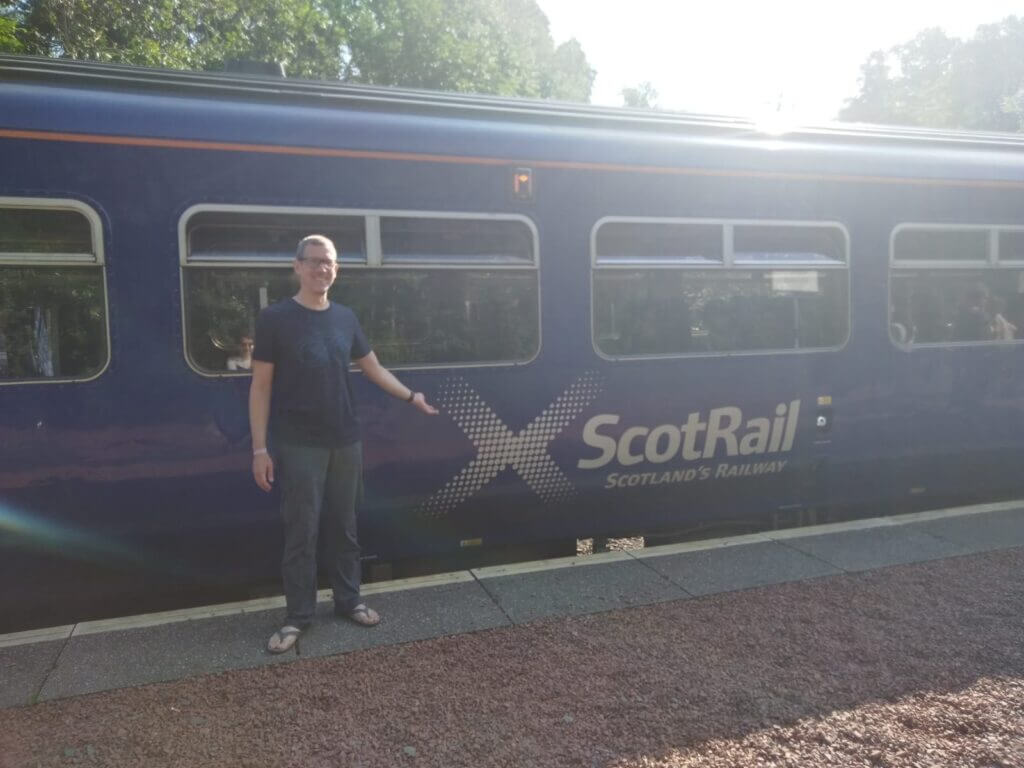 Ingo Steinke vor einem blauen Eisenbahnwaggon mit dem Logo der  ScotRail (Schottlands Eisenbahn) auf einem sonnigen Bahnsteig in den schottischen Highlands.