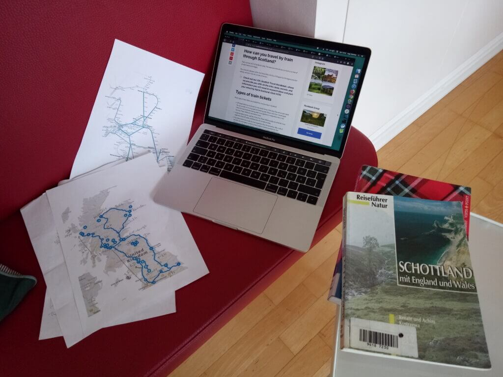 Travel guide books, printed train routes, and a laptop showing travel blogs on a red coffee house bench