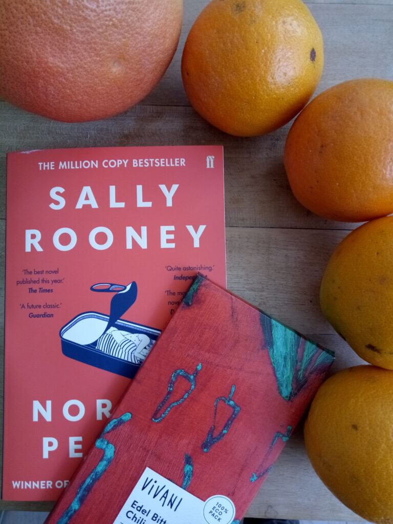 Rotes Cover von Sally Rooney's Normal People mit farblich passender Vivani-Schokolade und Orangen