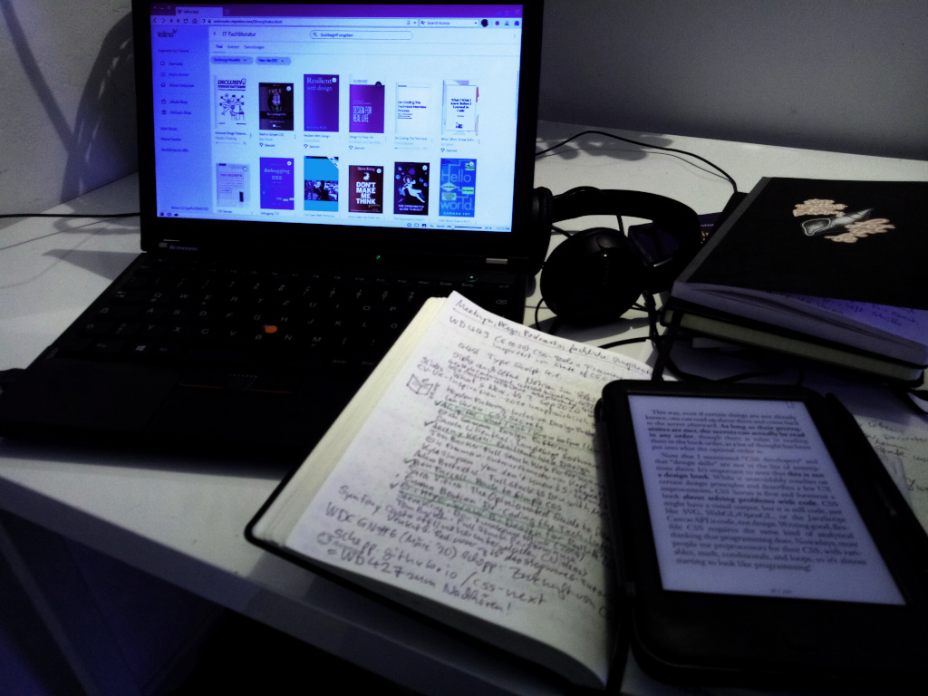 Schwarzes ThinkPad-Laptop mit Tolino-Webreader, schwarzes Notizbuch, Kopfhörer und Fachbücher auf einem schreibtisch