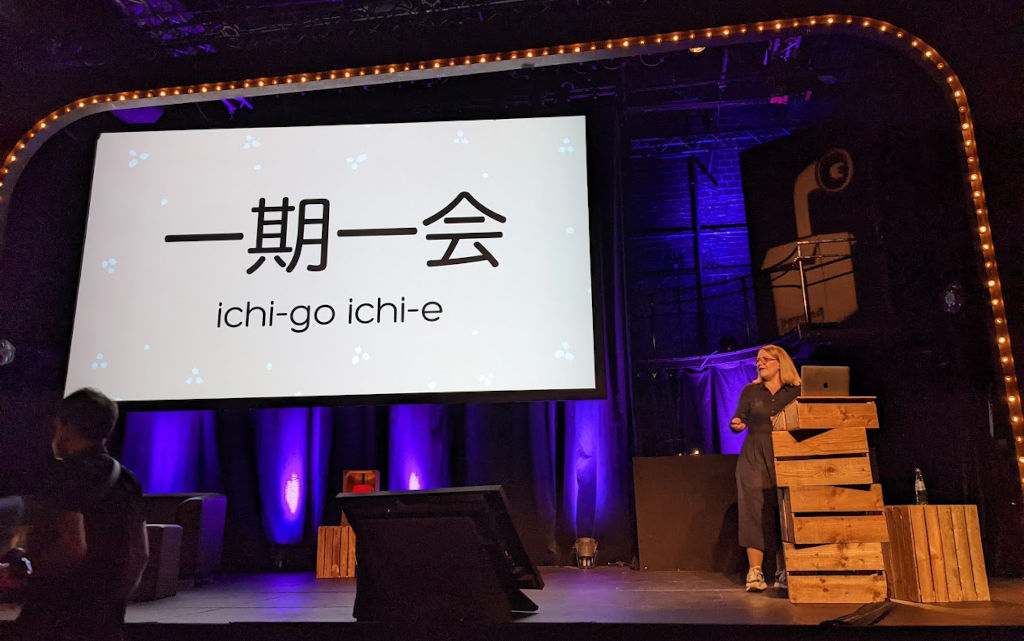 Stage screen showing the Japanese characters and pronunciation of ichi-go ichi-e and a woman presenting at a makeshift desk made out of wooden boxes