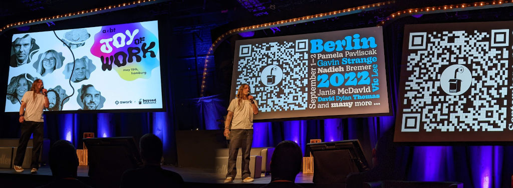 Marc Thiele, a man with long hair dressed in a casual t-shirt, in front of screens showing the announcements for the upcoming events in Hamburg and Berlin, including a QR code linking to the event page