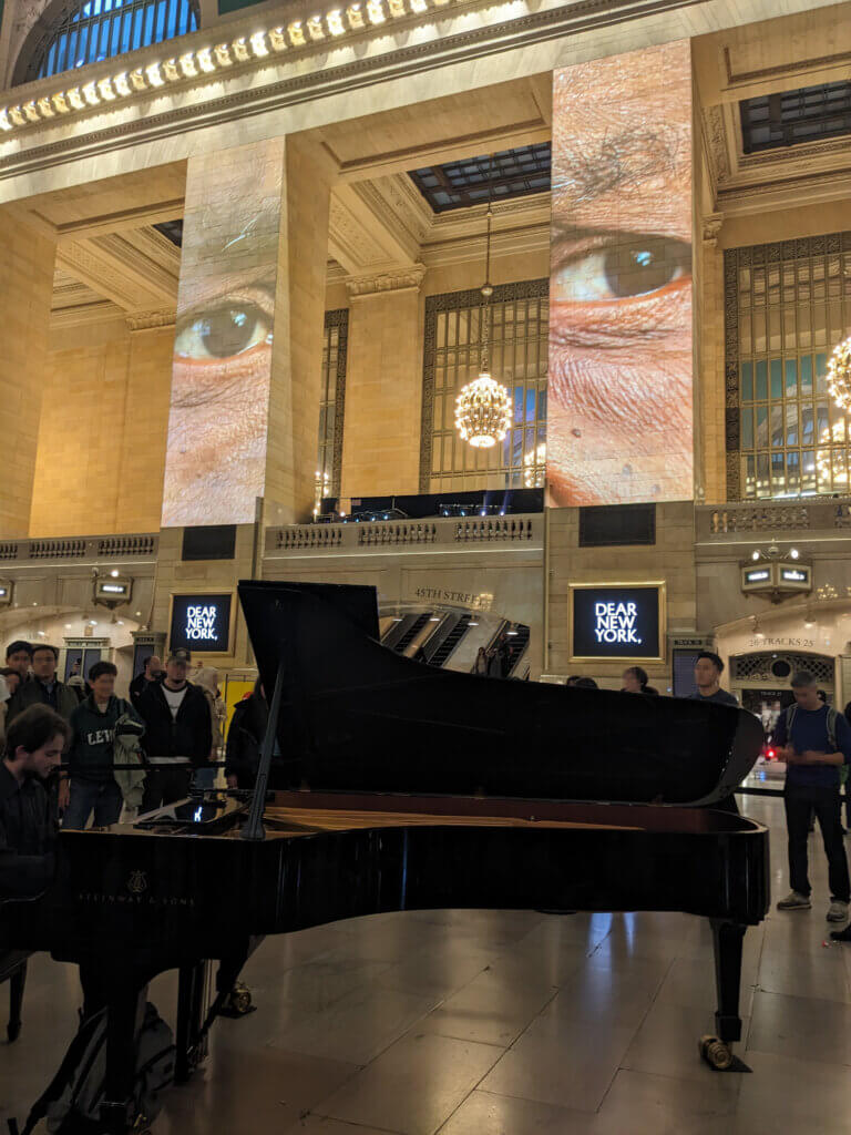 Piano player in Grand Central station below a projection of huge human eyes and a sign that says Dear New York