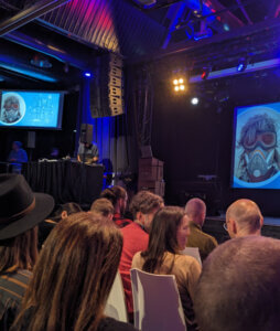 people discussing in the audience, looking at a picture of a masked speaker